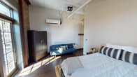 Located at 818 Georgia Ave in downtown Chattanooga, Tennessee, this studio unit blends industrial character with modern comfort. The open-plan living area showcases soaring ceilings with exposed ductwork and conduit, lending an authentic loft-style aesthetic. Rich, dark hardwood floors run throughout the space, grounding the industrial elements with warmth.

Exposed brick walls dominate one end of the studio, framing large, dark-trimmed windows that flood the interior with generous natural light and offer views of the surrounding urban streetscape. A private balcony door provides direct outdoor access. The sleeping and living areas flow seamlessly together, with the layout accommodating a full bed, a futon sofa, and a wardrobe armoire.

A compact kitchenette is neatly integrated into the space, featuring wall-mounted cabinetry, an under-mounted stainless steel sink, and countertop workspace — providing essential preparation amenities. A dedicated desk area positioned against the brick wall creates a functional workspace bathed in natural light.

The private bathroom is finished with tile flooring, a vanity with under-cabinet storage, a large mirror, and a shower-tub combination. A wall-mounted mini-split system ensures efficient climate control throughout. This unit offers a distinctive urban living experience rooted in Chattanooga's architectural heritage.