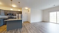This single-floor residence offers a thoughtfully designed open-concept layout that seamlessly connects the kitchen and living area. The kitchen is a standout feature, showcasing dark gray cabinetry, granite-style countertops, stainless steel appliances including a refrigerator, range, microwave, and dishwasher, and a peninsula with pendant lighting overhead. The adjoining living area is bright and open, finished with durable wood-look plank flooring that extends throughout the main living spaces, creating a cohesive and clean aesthetic. A dedicated entryway with a white panel door provides a welcoming transition into the home. The bedroom is carpeted for comfort and includes a spacious walk-in closet, providing generous storage. Two bathrooms feature matching gray vanities with white countertops, large mirrors, and tile-style flooring, offering a clean, modern finish. The neutral white walls throughout the unit provide a versatile backdrop, while the consistent finishes across rooms create a unified, contemporary feel. Natural light filters in through windows, enhancing the open and airy atmosphere of the space. Overall, this residence combines functional design with modern finishes, making it a comfortable and practical living environment.