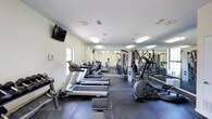 This fitness center offers a comprehensive workout environment designed to accommodate a wide range of exercise routines. The main workout area is generously proportioned, featuring a row of treadmills and elliptical machines lined along one side, ideal for cardio training. Full-length mirrors span an entire wall, enhancing the sense of space and allowing users to monitor their form during exercise. A multi-station cable machine and additional strength training equipment are positioned along the mirrored wall, providing options for a thorough resistance workout. The flooring throughout is a durable, padded rubber tile surface, providing both safety and comfort underfoot. Overhead, recessed fluorescent lighting ensures the space is well-lit at all times, complemented by natural light filtering in through windows. A dedicated free-weight area houses a tiered dumbbell rack with a full range of weights alongside an adjustable workout bench, creating a focused zone for strength training. A separate alcove provides a wall-mounted water fountain and hand sanitizer dispenser for convenience, along with a mini trampoline for low-impact exercise. The walls are finished in a clean, neutral tone, contributing to the bright and motivating ambiance of the facility.