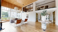 Located at 1 Cemetery St in Norwalk, CT, this thoughtfully designed single-floor space makes an immediate impression with its striking architectural details and welcoming ambiance. The open living area is anchored by soaring double-height ceilings clad in warm natural wood, creating a sense of grandeur that is both refined and inviting. Expansive floor-to-ceiling windows flood the interior with abundant natural light while framing lush outdoor views of the surrounding greenery and streetscape. Wide-plank hardwood flooring runs throughout, lending warmth and continuity to the space. A dramatic entrance area features a statement marble pedestal table beneath a mezzanine-level overlook with sleek black metal railings, setting a sophisticated tone from the moment of arrival. Built-in open shelving units with a rich wood finish line the walls of the corridor, offering both functional storage and a curated design element. The living room is elegantly appointed with contemporary furnishings, including a plush sectional sofa, a minimalist low-profile coffee table, and iconic globe pendant lighting that complement the clean, modern aesthetic. A dedicated lounge and café-style seating area adds versatility to the layout. The overall atmosphere balances modern design with natural materials, resulting in a bright, airy, and highly functional space.
