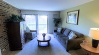 This single-floor space centers around a well-appointed living room designed for comfort and functionality. The living room is carpeted in a neutral tone that adds warmth underfoot and complements the light-colored walls. A large window fitted with vertical blinds allows generous natural light to fill the room, while offering a view of a balcony and surrounding greenery outdoors. An in-wall air conditioning unit is positioned near the window, providing climate control convenience.

The room is furnished with a spacious grey sectional-style sofa accented with decorative pillows, paired with a matching patterned armchair for additional seating. A round wooden coffee table with a lower shelf serves as the central focal point of the seating arrangement. A tall indoor plant adds a natural, refreshing element to the corner of the room, while a framed artwork piece on the wall contributes a decorative touch. A wooden storage unit or entertainment console is positioned along one wall, offering practical storage. Overall, the space presents a comfortable and functional living environment with a welcoming ambiance.