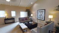 This single-story space makes a strong architectural impression through its vaulted ceilings accented by exposed decorative beams, which lend the living area a sense of openness and grandeur. The generous ceiling height draws the eye upward and contributes to an airy, spacious atmosphere throughout the main living area.

The living room is well-appointed and thoughtfully arranged, centered around a dark espresso-finished coffee table that anchors the seating arrangement. A neutral-toned sofa with burgundy and red accent pillows provides comfortable seating, complemented by a pair of tufted white ottomans that add a touch of elegance. A large framed artwork on the wall serves as a focal point, enhancing the refined aesthetic of the room.

Natural light filters in through multiple windows, including a prominent front-facing window near the entry door, brightening the warm beige walls and patterned area rug. Recessed lighting and a classic table lamp provide additional illumination. Tall indoor plants add organic texture and visual interest to the corners of the room. The overall ambiance is polished, welcoming, and well-balanced between comfort and style.