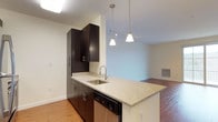 This 1-bedroom, 1-bathroom unit, designated as floor plan A1A, offers a thoughtfully arranged single-level layout that maximizes functionality within a compact footprint. The open-concept kitchen serves as a central feature, boasting rich dark espresso cabinetry that extends to the ceiling for ample storage. Granite countertops provide a neutral, speckled surface that contrasts elegantly with the dark cabinetry, while a stainless steel sink and dishwasher add a contemporary, practical touch. A peninsula-style counter extends into the living area, creating a natural divide between the kitchen and living spaces while offering potential for bar-style seating. Pendant lighting with frosted glass shades hangs above, complementing the track lighting visible in the adjoining living area. Hardwood-style flooring flows continuously throughout the space, contributing to a warm and cohesive aesthetic. The living area, visible from the kitchen, features neutral wall tones and appears well-lit, creating an inviting atmosphere. Clean lines, modern hardware, and quality finishes throughout reflect a contemporary design sensibility suited for comfortable everyday living.