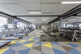 This expansive fitness center is designed to accommodate a comprehensive range of workout needs within a single, open-plan floor. The space is immediately distinguished by its bold, geometric-patterned carpet tiles in yellow, teal, and gray, adding a dynamic visual energy to the environment. Recessed linear lighting throughout the dropped ceiling ensures the entire floor is evenly and brightly illuminated.

The cardio zone runs along one side of the facility, featuring a long row of treadmills, ellipticals, StairMaster units, and stationary bikes positioned directly in front of large windows. These windows offer views of the surrounding urban streetscape and allow natural light to filter into the space. Wall-mounted televisions are strategically placed throughout this zone for entertainment during workouts.

The strength training area occupies the central and opposite portions of the floor, housing a robust selection of free weights, adjustable benches, cable machines, and multi-function cable systems. Full-length mirrors line sections of the walls, enhancing the sense of openness and supporting proper exercise form.

Entry to the facility is managed through a glass-walled, secured access corridor, providing a clean and professional transition into the gym. Overall, the space presents a well-organized, modern fitness environment suited for a wide variety of training programs.