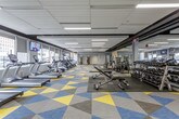 This expansive fitness center is designed to accommodate a comprehensive range of workout needs within a single, open-plan floor. The space is immediately distinguished by its bold, geometric-patterned carpet tiles in yellow, teal, and gray, adding a dynamic visual energy to the environment. Recessed linear lighting throughout the dropped ceiling ensures the entire floor is evenly and brightly illuminated.

The cardio zone runs along one side of the facility, featuring a long row of treadmills, ellipticals, StairMaster units, and stationary bikes positioned directly in front of large windows. These windows offer views of the surrounding urban streetscape and allow natural light to filter into the space. Wall-mounted televisions are strategically placed throughout this zone for entertainment during workouts.

The strength training area occupies the central and opposite portions of the floor, housing a robust selection of free weights, adjustable benches, cable machines, and multi-function cable systems. Full-length mirrors line sections of the walls, enhancing the sense of openness and supporting proper exercise form.

Entry to the facility is managed through a glass-walled, secured access corridor, providing a clean and professional transition into the gym. Overall, the space presents a well-organized, modern fitness environment suited for a wide variety of training programs.