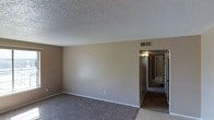 This single-floor space presents a practical and functional layout with a clean, understated aesthetic. The main living area is finished with wall-to-wall carpeting in a neutral tone, complementing the warm beige walls that run throughout. A textured popcorn ceiling adds a classic touch overhead, while standard white baseboards provide a clean border between the walls and floor.

Ventilation is handled through ceiling-mounted HVAC vents, ensuring climate control throughout the space. A centrally located hallway extends from the main living area, connecting to additional rooms further within the unit. The hallway is illuminated by a ceiling-mounted light fixture, and glimpses of natural light can be seen at the far end, suggesting windows in the adjoining rooms.

The overall color palette is cohesive and neutral, dominated by beige and taupe tones that lend a calm, move-in-ready atmosphere. The space is unfurnished, allowing prospective occupants a clear view of the layout and dimensions. The straightforward design prioritizes functionality, making it an adaptable space suited to a variety of furnishing and décor styles.