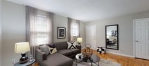 Located at 1076 Stuyvesant Ave in Irvington, NJ, this single-floor residence offers a thoughtfully laid-out living space with a neutral, modern aesthetic throughout. The living room is a welcoming area highlighted by warm parquet hardwood flooring, multiple windows dressed with sheer gray curtains that allow ample natural light, and a cohesive gray-toned color palette. The space comfortably accommodates a sectional sofa, area rug, and accent furnishings, creating an inviting atmosphere for relaxing or entertaining.

The kitchen is practical and well-equipped, featuring light wood cabinetry, dark countertops, black appliances including a gas range and microwave, and a sink beneath a window. White tile flooring runs through this area, which also accommodates a cozy dining nook with space for a small table and chairs. A door from the kitchen provides direct exterior access.

The bedroom is a serene retreat with parquet hardwood floors, multiple windows on two walls offering generous natural light, a ceiling fan fixture, and ample space for a full bedroom suite including a dresser and closet.

The full bathroom is clean and functional, finished with white tile flooring, white wainscoting, a vanity with under-sink storage, an oval mirror, and a shower-tub combination with a window for ventilation. Overall, the space presents a comfortable and move-in ready environment.
