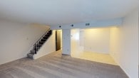 This two-story space offers a functional and straightforward layout designed to maximize usability across both levels. The main floor features a generously sized living area finished with wall-to-wall carpet in a neutral tone, providing a comfortable and cohesive foundation throughout the space. A textured white ceiling adds subtle dimension overhead, while multiple HVAC vents ensure consistent climate control throughout.

A staircase with a sleek black metal railing runs along one side of the main level, providing access to the upper floor while serving as a clean architectural accent within the space. Adjacent to the staircase, a doorway opens to what appears to be a kitchen or utility area, where a dark-finished appliance — likely a refrigerator — is visible, hinting at practical amenities nearby.

The main living area connects openly to an adjoining room finished with light-toned flooring, creating a sense of continuity and flow between spaces. The neutral color palette of soft whites and beiges throughout contributes to a bright, airy ambiance. Overall, the space presents a clean, move-in-ready environment well-suited for a variety of uses.