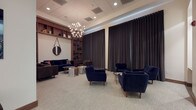 This refined single-floor space exudes a polished, contemporary ambiance crafted for comfort and professional sophistication. The main lounge area is generously proportioned, anchored by plush navy blue velvet sofas and armchairs arranged in thoughtful conversation groupings atop neutral, textured carpeting. A striking sputnik-style chandelier draws the eye upward, complementing the recessed lighting and a subtle LED cove that traces the ceiling perimeter.

Floor-to-ceiling dark drapery panels line the walls, lending a sense of drama and warmth, while rich walnut-toned built-in shelving units display books and decorative objects, adding depth and character to the interior. A leather armchair and a round wooden coffee table with curated reading material create an intimate nook framed by a circular hanging mirror.

An adjoining seating alcove features a pair of sleek dark armchairs set against clean white walls adorned with bold black-and-white botanical artwork, illuminated by a stylish tripod floor lamp. A separate transition area provides additional seating near a wall-mounted flat-screen television, a built-in desk nook, and a paneled wood entry door with transom window. Throughout, the space maintains a cohesive palette of navy, walnut, and warm neutrals, projecting a calm, upscale atmosphere suited to a premium lounge or professional reception environment.
