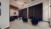 This refined single-floor space exudes a polished, contemporary ambiance crafted for comfort and professional sophistication. The main lounge area is generously proportioned, anchored by plush navy blue velvet sofas and armchairs arranged in thoughtful conversation groupings atop neutral, textured carpeting. A striking sputnik-style chandelier draws the eye upward, complementing the recessed lighting and a subtle LED cove that traces the ceiling perimeter.

Floor-to-ceiling dark drapery panels line the walls, lending a sense of drama and warmth, while rich walnut-toned built-in shelving units display books and decorative objects, adding depth and character to the interior. A leather armchair and a round wooden coffee table with curated reading material create an intimate nook framed by a circular hanging mirror.

An adjoining seating alcove features a pair of sleek dark armchairs set against clean white walls adorned with bold black-and-white botanical artwork, illuminated by a stylish tripod floor lamp. A separate transition area provides additional seating near a wall-mounted flat-screen television, a built-in desk nook, and a paneled wood entry door with transom window. Throughout, the space maintains a cohesive palette of navy, walnut, and warm neutrals, projecting a calm, upscale atmosphere suited to a premium lounge or professional reception environment.