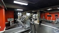 This fitness center is a dedicated, single-floor workout facility designed with functionality and efficiency in mind. The space features a selection of cardio equipment, including treadmills and elliptical machines, arranged to allow comfortable movement between stations. Full-length mirrored walls line the room, visually expanding the space and providing users with proper form feedback during exercise. The dark ceiling, contrasted by bold red accent walls, lends the gym a dynamic and energetic atmosphere. Overhead fluorescent lighting ensures the space is well-lit throughout. The floor is finished with durable rubber gym flooring, offering both safety and comfort during high-impact activities. The layout is practical and open, making efficient use of the available floor space. Exposed structural elements on the ceiling contribute to an industrial aesthetic that complements the gym's no-frills, performance-focused design. Overall, this fitness center provides a compact yet comprehensive environment suited for cardiovascular training and general fitness routines.