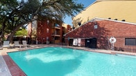 This outdoor pool area serves as a central amenity within a multi-story residential community, offering a well-maintained and inviting retreat for relaxation and recreation. The centerpiece is a generously sized swimming pool with clear, turquoise water, complemented by an adjacent spa, providing options for both active swimming and leisurely soaking. The expansive concrete sundeck surrounding the pool is furnished with lounge chairs and umbrella-shaded seating, offering ample space for sunbathing and outdoor gatherings. The area is enclosed by a wrought iron fence, ensuring both security and an open, airy aesthetic. Mature trees provide natural shade throughout the space, creating a comfortable environment even on sunny days. The pool area is framed by multi-story brick and wood-sided residential buildings, lending the space a cohesive and established community feel. Access to the pool is provided through a dedicated entrance with dark wood-framed glass doors, leading from a covered exterior vestibule. Posted pool rules are prominently displayed at the entry point, reflecting attention to community standards. Overall, this pool area combines functional amenities with a pleasant outdoor ambiance, making it a desirable shared space within the residential community.