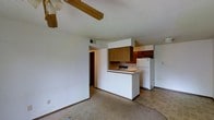 This single-floor space offers a functional and open layout that connects the living area directly to the kitchen, creating a cohesive and practical environment. The living area features carpeted flooring, neutral white walls, and a ceiling fan with exposed wooden beam detailing set against a textured popcorn ceiling, lending a casual and comfortable character to the space.

The kitchen is defined by a peninsula-style breakfast bar that serves as a natural divider between the two areas. Warm wood-toned upper and lower cabinetry provides ample storage, complemented by a built-in cooktop and a full-size white refrigerator. The kitchen floor transitions to tile, distinguishing it clearly from the carpeted living space.

A hallway visible at the rear of the unit leads to additional rooms, suggesting a well-organized floor plan that separates the private and shared living areas. Overhead ventilation is provided via ceiling-mounted HVAC vents, ensuring climate control throughout. The overall ambiance is straightforward and utilitarian, with a neutral color palette that offers a blank canvas for personalization. The layout maximizes the usability of the available space, making it well-suited for everyday living.