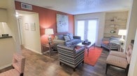 This single-story space centers around a well-appointed living area that combines contemporary design elements with a warm, inviting atmosphere. A striking terracotta-red accent wall serves as the focal point of the room, complemented by neutral surrounding walls and a textured popcorn ceiling that adds depth overhead.

The living area is furnished with a grey sectional sofa adorned with patterned throw pillows, alongside a bold chevron-print armchair that adds a dynamic visual contrast. A sleek glass-and-chrome coffee table anchors the seating arrangement, while a matching side table with a classic table lamp adds warmth to the space. An area rug in a rich red-orange tone defines the seating zone atop the dark hardwood-style flooring, which runs throughout and adds a grounding, cohesive quality to the room.

Large French-style patio doors dominate one wall, fitted with white horizontal blinds that allow for adjustable natural light. A built-in shelving unit is partially visible near the entrance, offering practical storage. A framed art piece above the sofa and wall art on the accent wall contribute to the curated, modern aesthetic of the space.