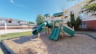 This outdoor community space offers a variety of amenities set against a backdrop of multi-story residential buildings constructed with classic brick and siding exteriors. The centerpiece of the grounds is a substantial multi-level playground structure finished in green and wood tones, featuring enclosed tube slides, open slides, climbing elements, and elevated platforms. The playground sits within a generously sized wood chip safety surface, clearly bordered and well-maintained to ensure safe play.

Adjacent to the playground is an open, grassy lawn area that provides additional space for outdoor activities. A sand or gravel recreational court is also visible nearby, offering further options for outdoor enjoyment. The grounds are landscaped with mature trees, manicured shrubs, and maintained turf, contributing to a green and pleasant atmosphere throughout.

A small single-story utility or amenity building is present on the property, constructed with a matching brick lower facade and siding upper section beneath a simple pitched roof. A black metal bench is positioned just outside, creating a quiet spot for residents to sit outdoors.

Ample paved parking is available on-site, with bike racks also provided near the buildings. The overall environment is clean, open, and well-kept, offering residents practical outdoor amenities in a comfortable community setting.