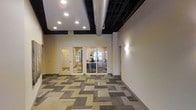 This single-floor commercial space combines industrial and modern design elements to create a functional and visually engaging environment. The open corridor or reception area immediately draws attention with its bold geometric carpet tiles, which feature an interlocking pattern of neutral tones including beige, gray, and taupe, adding a dynamic quality to the flooring throughout the space.

The ceiling design is a notable architectural feature, showcasing a combination of a recessed, framed drop section with embedded recessed lighting alongside an exposed dark-painted structure with visible silver ductwork. This contrast between the refined ceiling tray and the raw industrial elements above gives the space a layered, contemporary character.

Interior glass-paneled walls and doors provide visual connectivity between the corridor and the adjoining offices or workrooms, allowing natural light to filter through while maintaining defined zones. Wall-mounted sconce lighting adds a warm accent along the neutral-toned walls. The offices visible through the glass appear furnished and operational, suggesting an active professional or administrative environment. Overall, the space presents a clean, well-lit, and purposefully designed commercial interior.