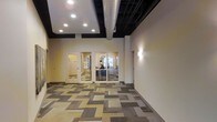 This single-floor commercial space combines industrial and modern design elements to create a functional and visually engaging environment. The open corridor or reception area immediately draws attention with its bold geometric carpet tiles, which feature an interlocking pattern of neutral tones including beige, gray, and taupe, adding a dynamic quality to the flooring throughout the space.

The ceiling design is a notable architectural feature, showcasing a combination of a recessed, framed drop section with embedded recessed lighting alongside an exposed dark-painted structure with visible silver ductwork. This contrast between the refined ceiling tray and the raw industrial elements above gives the space a layered, contemporary character.

Interior glass-paneled walls and doors provide visual connectivity between the corridor and the adjoining offices or workrooms, allowing natural light to filter through while maintaining defined zones. Wall-mounted sconce lighting adds a warm accent along the neutral-toned walls. The offices visible through the glass appear furnished and operational, suggesting an active professional or administrative environment. Overall, the space presents a clean, well-lit, and purposefully designed commercial interior.