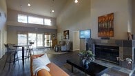 This single-story space functions as a welcoming common area or clubhouse, distinguished by its dramatic vaulted ceilings that lend an expansive, airy quality to the interior. The open layout is anchored by a sleek, floor-to-ceiling stone fireplace paired with a wall-mounted television, creating a natural focal point for the main lounge area. Rich, dark hardwood-style flooring runs throughout, adding warmth and cohesion to the space.

The seating area is furnished with a tan leather sofa, patterned accent chairs, and a low-profile coffee table set atop a textured area rug, offering a comfortable and inviting atmosphere. Pendant lighting and wall-mounted sconces complement the natural light that pours in through large windows positioned high on the walls and a set of French doors at the rear, which open to an outdoor area and frame views of surrounding greenery.

On one side, a kitchenette or bar-style counter is partially visible, suggesting additional functionality. Decorative elements including a bold autumn landscape painting and potted plants add color and life to the neutral-toned walls. The overall ambiance is modern, open, and well-appointed, designed to encourage gathering and relaxation.