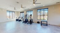 Located at 305 Prine Pl in Charlotte, NC, this contemporary fitness center offers a well-appointed, single-floor layout designed to support a full range of workout routines. The space is bathed in natural light through an abundance of large, black-framed windows that frame views of the surrounding greenery and neighboring community, creating an open and energizing atmosphere.

The main cardio area is generously proportioned, housing multiple elliptical machines, stationary bikes, and treadmills arranged to maximize usability and comfort. A full-wall mirror in the treadmill section enhances the sense of space while allowing users to monitor their form. Polished concrete-style flooring runs throughout, providing a durable and sleek surface suited to high-traffic fitness use.

Overhead, stylish large-blade ceiling fans with brushed gold accents contribute both to the modern aesthetic and to air circulation across the space. Recessed lighting supplements the abundant natural light, ensuring the facility remains bright and welcoming at any hour. Additional fitness accessories, including stability balls and free weights, are available throughout.

The open floor plan allows for flexible use, with ample room for stretching and functional training. The overall design balances clean, neutral tones with modern fixtures, delivering a motivating and comfortable environment for fitness enthusiasts in Charlotte.