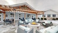 Located at 38 Westgate Dr. in Dover, New Hampshire, this single-level outdoor amenity space is a thoughtfully designed community patio built for both relaxation and entertainment. The centerpiece of the space is a striking timber-frame structure with an exposed wood gable roof, which shelters a generous covered lounge area beneath pendant lighting. The warm natural wood of the timber framing contrasts beautifully against the crisp white lap siding of the adjacent building, creating an inviting, coastal-influenced aesthetic.

The patio is finished with large-format stone pavers that extend throughout the space, providing a clean and durable foundation. A circular stone fire pit serves as a natural gathering point, surrounded by wicker lounge chairs and sofas upholstered in soft aqua cushions. An adjacent pergola extends the outdoor living area, offering an open-air dining zone furnished with a full table and seating for multiple guests.

A built-in outdoor kitchen station features dual stainless steel grills set into a stone surround, making the space well-equipped for outdoor cooking and entertaining. The surrounding landscape includes manicured shrubs along the building's stone-accented foundation, with a backdrop of mature trees and open views of the broader property. This outdoor space offers a well-rounded and cohesive environment suited for community gathering year-round.