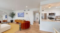 This single-floor residence offers a cohesive and stylish layout across a well-utilized footprint. The open-concept living room and kitchen area serves as the heart of the home, featuring light hardwood flooring, recessed lighting, and crisp white walls that create a bright, airy ambiance. The living room is generously proportioned, offering ample space for seating arrangements, and flows seamlessly into the kitchen. The kitchen is designed in a galley configuration, outfitted with white shaker cabinetry, white quartz countertops, stainless steel appliances including a range and dishwasher, and a subway tile backsplash — combining functionality with clean aesthetics. The bedroom is a comfortable retreat, appointed with a ceiling fan, large windows allowing natural light, and light hardwood flooring. The bathroom features a white vanity with a quartz countertop, tiled flooring, and a shower-tub combination. A dedicated office provides a productive workspace with built-in wood bookshelves, French doors, a ceiling fan, and a distinctive checkered carpet. A connecting hallway ties the rooms together efficiently. The overall design balances mid-century modern warmth with contemporary clean lines, resulting in a polished and livable space.