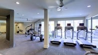 This fitness center is a thoughtfully designed workout space that offers everything needed for an effective cardio-focused exercise routine. The room is bright and inviting, with large windows lining the walls and allowing generous amounts of natural light to flood the interior, creating an open and energizing atmosphere. Views of the surrounding exterior add a pleasant outdoor connection while working out.

The space is furnished with a comprehensive selection of cardio equipment, including multiple treadmills, elliptical machines, and a stair climber, all arranged to allow comfortable movement throughout the room. Structural columns are integrated cleanly into the layout without disrupting the flow of the space. Overhead, ceiling fans are strategically placed to maintain air circulation and comfort during workouts, complementing the recessed lighting that ensures even, consistent illumination throughout. The flooring features a durable, patterned carpet designed to withstand heavy equipment and high foot traffic. The neutral wall tones and clean finishes give the space a modern, professional aesthetic. Overall, this fitness center strikes a balance between functionality and comfort, making it a practical and welcoming environment for daily exercise.