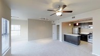 This single-floor space offers a clean, open-concept layout that connects the living area and kitchen in a seamless, functional design. The living area features plush carpeted flooring in a soft neutral tone, with smooth white ceilings and a five-blade ceiling fan with an integrated light fixture providing both comfort and illumination. Track lighting is also installed, offering flexible lighting options throughout the space. The walls are finished in a warm beige tone, creating a calm and inviting ambiance. A closet with louvered doors adds convenient built-in storage. The kitchen is defined by a raised bar-height counter with a light stone surface, complemented by sleek dark cabinetry that provides a modern contrast. Pendant lights with deep red accents hang above the counter, adding a stylish focal point. A stainless steel refrigerator is visible in the kitchen, reinforcing the contemporary aesthetic. The overall design balances practicality with modern style, featuring thoughtful details such as recessed HVAC vents and a well-considered flow between living and culinary spaces. The neutral palette and open layout make the space highly adaptable to a variety of interior design preferences.