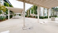 Located at 11850 Florence Ave in Santa Fe Springs, California, this generously sized outdoor communal space serves as a central gathering area within a residential community. Spanning a substantial footprint on a single level, the space is thoughtfully designed to encourage outdoor living and social interaction.

At the heart of the area, a series of pergola-covered concrete patios provide shade and structure, supported by clean-lined columns and slatted overhead trellises that filter natural light. Concrete picnic tables offer comfortable seating for dining and socializing, while multiple charcoal BBQ grills are conveniently positioned nearby for outdoor cooking and gatherings.

Well-maintained landscaping runs throughout the space, featuring a mix of ornamental trees, shrubs, succulents, and ground cover plantings set against mulched beds. Manicured green lawns provide open recreational space, adding a refreshing natural contrast to the smooth concrete pathways and walkways that connect each area.

A children's playground, equipped with a colorful play structure including slides and climbing elements, is situated on a sandy base within clear view of the pergola areas, making supervision easy. Decorative street-style lamp posts line the pathways, enhancing both the aesthetic and functionality of the space after dark. The surrounding residential buildings, finished in white stucco, frame the courtyard and contribute to its welcoming, community-centered atmosphere.
