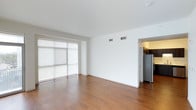 This single-floor space showcases a clean, contemporary design with a minimalist aesthetic throughout. The main living area features warm-toned hardwood flooring that extends seamlessly across the open layout, complemented by crisp white walls and a smooth white ceiling that enhances the sense of brightness and openness. Large floor-to-ceiling sliding glass doors dominate one wall of the living space, fitted with sheer roller shades that diffuse natural light softly into the room while maintaining privacy. The generous glazing creates a strong connection to the outside and contributes to the airy, open feel of the interior. Overhead, centrally placed HVAC vents are neatly integrated into the ceiling, ensuring efficient climate control without disrupting the clean lines of the space. Adjacent to the main living area, an opening leads directly into the kitchen, where a stainless steel refrigerator is visible, hinting at modern appliances throughout. The overall ambiance is bright, neutral, and move-in ready, offering a versatile canvas well-suited for a variety of interior styles and furnishing arrangements.