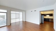 This single-floor space showcases a clean, contemporary design with a minimalist aesthetic throughout. The main living area features warm-toned hardwood flooring that extends seamlessly across the open layout, complemented by crisp white walls and a smooth white ceiling that enhances the sense of brightness and openness. Large floor-to-ceiling sliding glass doors dominate one wall of the living space, fitted with sheer roller shades that diffuse natural light softly into the room while maintaining privacy. The generous glazing creates a strong connection to the outside and contributes to the airy, open feel of the interior. Overhead, centrally placed HVAC vents are neatly integrated into the ceiling, ensuring efficient climate control without disrupting the clean lines of the space. Adjacent to the main living area, an opening leads directly into the kitchen, where a stainless steel refrigerator is visible, hinting at modern appliances throughout. The overall ambiance is bright, neutral, and move-in ready, offering a versatile canvas well-suited for a variety of interior styles and furnishing arrangements.