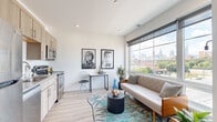 This single-floor residence is thoughtfully designed with a contemporary aesthetic, combining clean lines, light-toned finishes, and an open layout that maximizes the sense of space throughout. Light hardwood flooring runs continuously through the main living areas, complementing the bright white walls and recessed lighting overhead.

The living area serves as the centerpiece of the unit, anchored by a large picture window that frames impressive city skyline views, flooding the space with natural light. A compact desk area tucked along one wall provides a functional workspace without interrupting the flow of the room.

The kitchen is positioned adjacent to the living space and features light wood cabinetry, white subway tile backsplash, and a full suite of stainless steel appliances, including a refrigerator, range, microwave, and dishwasher — offering a well-appointed cooking environment.

The bedroom is a calm, white-walled retreat with a notable clerestory window positioned near the ceiling, drawing in additional natural light. A closet provides practical storage within the room.

The bathroom is clean and functional, finished with tile flooring and a full tub-shower combination, a single vanity with light wood cabinetry, and crisp white walls throughout.