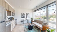 This single-floor residence is thoughtfully designed with a contemporary aesthetic, combining clean lines, light-toned finishes, and an open layout that maximizes the sense of space throughout. Light hardwood flooring runs continuously through the main living areas, complementing the bright white walls and recessed lighting overhead.

The living area serves as the centerpiece of the unit, anchored by a large picture window that frames impressive city skyline views, flooding the space with natural light. A compact desk area tucked along one wall provides a functional workspace without interrupting the flow of the room.

The kitchen is positioned adjacent to the living space and features light wood cabinetry, white subway tile backsplash, and a full suite of stainless steel appliances, including a refrigerator, range, microwave, and dishwasher — offering a well-appointed cooking environment.

The bedroom is a calm, white-walled retreat with a notable clerestory window positioned near the ceiling, drawing in additional natural light. A closet provides practical storage within the room.

The bathroom is clean and functional, finished with tile flooring and a full tub-shower combination, a single vanity with light wood cabinetry, and crisp white walls throughout.