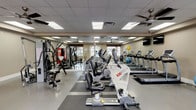This fitness center offers a comprehensive workout environment designed to accommodate a wide range of exercise routines. The space is outfitted with a strong selection of cardio equipment, including treadmills, elliptical trainers, and stationary bikes, all arranged to maximize usability and flow throughout the room. A dedicated strength training station anchors one side of the facility, complemented by a rack of free weights and medicine balls for functional training. The flooring features durable rubber matting in workout zones, providing both safety and comfort during exercise. Large windows along the perimeter allow natural light to filter in, creating an open and energizing atmosphere. Multiple ceiling fans are mounted overhead, ensuring proper air circulation throughout the space. A dropped ceiling with recessed panel lighting provides consistent, even illumination across the entire facility. Wall-mounted mirrors and flat-screen televisions add to the functionality and comfort of the environment. The neutral-toned walls and clean layout give the space a professional yet welcoming feel, making it suitable for a variety of fitness levels and workout styles.