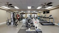 This fitness center offers a comprehensive workout environment designed to accommodate a wide range of exercise routines. The space is outfitted with a strong selection of cardio equipment, including treadmills, elliptical trainers, and stationary bikes, all arranged to maximize usability and flow throughout the room. A dedicated strength training station anchors one side of the facility, complemented by a rack of free weights and medicine balls for functional training. The flooring features durable rubber matting in workout zones, providing both safety and comfort during exercise. Large windows along the perimeter allow natural light to filter in, creating an open and energizing atmosphere. Multiple ceiling fans are mounted overhead, ensuring proper air circulation throughout the space. A dropped ceiling with recessed panel lighting provides consistent, even illumination across the entire facility. Wall-mounted mirrors and flat-screen televisions add to the functionality and comfort of the environment. The neutral-toned walls and clean layout give the space a professional yet welcoming feel, making it suitable for a variety of fitness levels and workout styles.