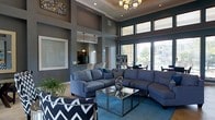 This single-floor space makes a striking first impression with its dramatically high ceilings and bold, contemporary color palette of deep blues and cool greys. The main living area is generously proportioned, anchored by a large sectional sofa in navy blue, complemented by a chevron-patterned accent chair and a sleek glass and metal coffee table. An elegant chandelier hangs overhead, adding warmth to the otherwise modern aesthetic, while recessed lighting provides additional illumination throughout.

Floor-to-ceiling windows along one wall flood the space with natural light and offer views of the surrounding exterior, enhancing the open, airy feel. Tall structural columns frame the room, adding architectural definition. A teal-blue carpet grounds the seating area, tying the color scheme together cohesively.

Visible beyond the main living space is a kitchen or service area, distinguished by pendant lighting and a pass-through counter. Additional seating and table arrangements near the windows suggest a flexible, multi-use layout. Overall, the space exudes a polished, modern ambiance suited for both relaxed gatherings and more formal settings.