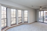 This single-floor space is thoughtfully laid out across several distinct rooms, offering a functional and comfortable living environment. The spacious living area is a standout feature, anchored by a large bay window arrangement that frames outdoor views and fills the room with natural light. Soft, neutral-toned carpet and a textured ceiling contribute to the room's warm, understated character.

An arched entryway connects the living area to an additional carpeted room, which features its own window arrangement and provides flexible use as a secondary living space or den. Both carpeted areas are finished with warm wood baseboards that complement the overall neutral palette.

The galley-style kitchen is fitted with oak cabinetry providing generous storage on both sides, white countertops, wood-look flooring, and black appliances including a refrigerator, electric range, dishwasher, and overhead ventilation hood. A circular window above the sink adds a distinctive architectural detail.

The bedroom offers ample storage with two sliding-door closets and an additional standard door, all trimmed in warm oak tones, with plush carpeting and a window for natural light.

The bathroom is finished with wood-look flooring and includes a full tub-shower combination, toilet, and vanity, maintaining a clean, practical design consistent with the rest of the space.