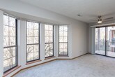This single-floor space is thoughtfully laid out across several distinct rooms, offering a functional and comfortable living environment. The spacious living area is a standout feature, anchored by a large bay window arrangement that frames outdoor views and fills the room with natural light. Soft, neutral-toned carpet and a textured ceiling contribute to the room's warm, understated character.

An arched entryway connects the living area to an additional carpeted room, which features its own window arrangement and provides flexible use as a secondary living space or den. Both carpeted areas are finished with warm wood baseboards that complement the overall neutral palette.

The galley-style kitchen is fitted with oak cabinetry providing generous storage on both sides, white countertops, wood-look flooring, and black appliances including a refrigerator, electric range, dishwasher, and overhead ventilation hood. A circular window above the sink adds a distinctive architectural detail.

The bedroom offers ample storage with two sliding-door closets and an additional standard door, all trimmed in warm oak tones, with plush carpeting and a window for natural light.

The bathroom is finished with wood-look flooring and includes a full tub-shower combination, toilet, and vanity, maintaining a clean, practical design consistent with the rest of the space.