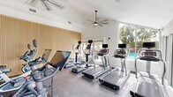 Located at 1130 E 3rd in Oakland, CA, this fitness center is a thoughtfully designed single-floor facility comprising two exercise rooms connected by a hallway. The space delivers a complete workout environment with a clean, modern aesthetic throughout.

The first exercise room is bright and open, benefiting from abundant natural light streaming through large sliding glass doors that lead to an outdoor terrace. Treadmills and stationary cycling bikes are arranged across durable rubber flooring, while the expansive windows offer views of the surrounding greenery and skyline — creating an energizing atmosphere for cardio training.

The second exercise room features bold navy blue accent walls paired with full-length mirrored panels that visually expand the space. This room is dedicated to strength training, housing a dumbbell rack, weight benches, cable machines, and additional resistance equipment. Recessed lighting and a wall-mounted air conditioning unit ensure comfort during intense workouts.

A third area, visible through mirroring and the layout, incorporates more cardio machines — including ellipticals and upright bikes — set against a striking slatted wood accent wall that adds warmth and texture to the otherwise crisp interior. The connecting hallway provides seamless flow between areas. Together, the rooms offer a well-rounded, professionally outfitted fitness facility suitable for a wide range of training needs.