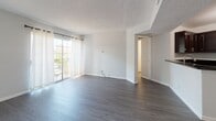 This single-floor unit presents a clean, neutral interior characterized by consistent dark hardwood-style flooring throughout most of the living and sleeping areas, complemented by crisp white baseboards and light gray walls. The entry area welcomes residents with a pendant light fixture overhead and direct access to the main living space.

The spacious living room is a standout feature, offering sliding glass doors framed by sheer white curtains that allow abundant natural light and provide views of outdoor greenery, including a visible palm tree. A textured popcorn ceiling adds a classic touch throughout the unit.

The kitchen is well-appointed with dark espresso cabinetry, dark countertops, a stainless steel sink, and a full suite of stainless steel appliances including a gas range, over-the-range microwave, and refrigerator. Light-toned flooring in the kitchen contrasts effectively with the darker cabinetry.

The bedrooms are generously sized, each featuring windows with blinds that bring in natural light and views of the surrounding landscape. The bathroom includes a full tub and shower combination with white tile surround, chrome fixtures, a toilet, and a vanity with white cabinetry.