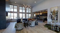 This single-floor space makes a striking impression with its dramatic double-height ceilings and expansive floor-to-ceiling windows that flood the interior with abundant natural light. The open-concept layout connects a sophisticated lounge area to a fully equipped kitchen and bar, creating a cohesive and versatile environment.

The lounge area is anchored by an eclectic mix of seating, including a tufted navy sofa, a rich cognac leather chair, and a pair of patterned swivel chairs featuring a bold tree-branch motif. A multi-bulb chandelier with an industrial-modern aesthetic serves as a focal point overhead, complementing the warm tones of the light hardwood flooring that runs throughout the space.

Transitioning toward the kitchen and bar area, dark cabinetry and a marble-topped island provide a refined contrast to the lighter tones of the lounge. Woven globe pendant lights hang above the bar seating, adding warmth and texture. The overall ambiance is contemporary and curated, balancing bold design elements with an inviting, open atmosphere suitable for both social gatherings and everyday use.