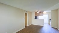 This single-floor residence features an open-concept living area finished with durable wood-style laminate flooring that extends throughout the main space. The living room is generously proportioned, with neutral beige walls and a textured popcorn ceiling overhead. A ceiling fan with integrated lighting is centrally positioned, providing both air circulation and ambient illumination.

A doorway off the main living area leads to an adjoining room, likely a hallway or additional living space. Toward the far end of the room, a peninsula-style counter separates the living area from the kitchen, offering a pass-through design that enhances the open feel of the layout. The kitchen features dark countertops and upper cabinetry in a warm wood tone.

Natural light enters through a large window fitted with vertical blinds along the far wall, brightening the interior and contributing to an airy atmosphere. A centrally located HVAC vent in the ceiling ensures climate control throughout the space. White baseboards frame the perimeter of the room cleanly, complementing the neutral color palette. Overall, the space presents a clean, functional layout suited for everyday living.