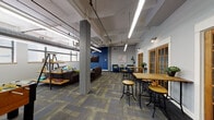 This single-floor space showcases a well-executed industrial-modern design, combining exposed structural elements with comfortable, thoughtfully arranged furnishings. The soaring ceilings reveal exposed ductwork, conduit, and linear LED lighting suspended from white-painted beams, lending an open, loft-like character to the interior. Concrete support columns punctuate the floor plan, reinforcing the industrial aesthetic while defining distinct functional zones across the carpeted floor.

The central lounge area features a generously sized sectional and sofa arrangement in dark leather, surrounding a rustic wooden coffee table with industrial caster wheels. Colorful accent pillows in green and teal introduce a lively contrast. Adjacent to this, a dedicated seating nook hosts two navy blue armchairs with yellow accent pillows set against a bold blue accent wall, accompanied by a wall-mounted flat-screen television and curated artwork.

Along the perimeter, tall wooden bar tables paired with adjustable industrial stools provide casual collaborative or individual work surfaces. A row of warm-toned wood-framed French doors lines one wall, allowing natural light to filter through while maintaining visual connectivity between zones. Additional details include potted greenery, a foosball table visible in the background, and eclectic artwork, all contributing to an inviting and dynamic atmosphere.