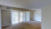 This single-floor space presents a clean, understated interior highlighted by a generously sized living area. The room is anchored by a wide wall of sliding glass doors dressed with full-length vertical blinds, allowing abundant natural light to filter in while offering a view of the greenery outside. The neutral, light-toned walls complement the wall-to-wall carpeting, which provides a soft, understated foundation throughout the space. A subtle architectural ledge runs along the upper portion of the exterior wall, adding a modest structural detail to the otherwise straightforward design. Ceiling-mounted fixtures and integrated wall vents indicate climate control infrastructure built directly into the unit. The open layout of the living area offers flexibility in furniture arrangement, making it adaptable to various lifestyle needs. Overall, the space maintains a simple, functional aesthetic with an emphasis on natural light and an open feel, creating a calm and neutral environment well-suited for personalization.