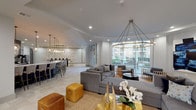 Located at 4220 San Felipe St in Houston's prestigious Galleria corridor, this sophisticated single-floor community clubroom offers an exceptional gathering space designed with contemporary elegance. The open-concept layout flows seamlessly between distinct functional zones, all unified by light-toned tile flooring and a refined neutral palette accented with gold and navy tones.

The main lounge area is anchored by a generous sectional sofa arrangement beneath a striking brass hoop chandelier with dramatic pendant detailing. Curated artwork and gold-accented décor add warmth and personality throughout. A dedicated conversation nook features navy velvet wingback chairs arranged around a central table, bathed in natural light from expansive floor-to-ceiling windows that frame views of the landscaped courtyard below.

The kitchen and bar area is outfitted with bold navy cabinetry, quartz countertops, and a dark subway tile backsplash accented by decorative round mirrors. Pendant lighting clusters illuminate the long island counter with bar seating, creating an ideal space for social gatherings. Appliances including a coffee station and microwave enhance everyday functionality.

Folding glass doors connect the interior to an adjacent lounge space, extending the versatility of the layout. Recessed lighting throughout ensures consistent illumination, while the abundant glazing fills the space with natural light and a strong visual connection to the outdoor amenity areas.