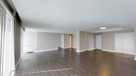 This single-floor space presents a generously sized, open-plan layout finished with rich, dark hardwood flooring that runs throughout, creating a cohesive and polished aesthetic. The expansive main living area is defined by smooth, light-coloured ceilings and clean, neutral grey walls accented by crisp white baseboards, lending the space a modern yet understated character. Natural light filters into the room, casting warm reflections across the flooring and enhancing the sense of openness.

Structural columns subtly divide the space, creating distinct zones within the open layout without compromising the overall sense of flow. Multiple doorways are visible from the main area, indicating access to additional rooms beyond, including what appears to be a corridor leading further into the unit and a dedicated entry door. A flush ceiling-mounted light fixture provides overhead illumination in one section, while the layout benefits from an abundance of ambient light throughout.

The space is presented in a clean, move-in ready condition, offering a neutral and versatile canvas suitable for a variety of interior configurations and design styles. The overall ambiance is calm, contemporary, and well-proportioned.