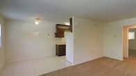 Located at 1521 Rolling Glen Drive in Wilmington, Delaware, this single-floor unit offers a straightforward and functional layout designed for comfortable everyday living. The living area features wall-to-wall carpeting that adds warmth underfoot, while light-toned walls keep the space feeling bright and open. A flush-mounted ceiling fixture provides overhead lighting throughout the main area. The kitchen is positioned adjacent to the living space and is defined by dark wood cabinetry that provides ample storage above and below the counter. A white refrigerator anchors one side of the kitchen, and the tile flooring in this area offers a clean, easy-to-maintain surface that contrasts with the carpeted living zone. A partial wall separates the two areas, creating a subtle sense of definition without fully closing off the space. A fire extinguisher is visibly mounted on the kitchen wall, reflecting attention to safety. The overall layout is compact yet practical, with neutral finishes throughout that offer a clean canvas for personalization. The single-floor design ensures convenient, step-free accessibility across the entire unit.