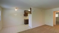 Located at 1521 Rolling Glen Drive in Wilmington, Delaware, this single-floor unit offers a straightforward and functional layout designed for comfortable everyday living. The living area features wall-to-wall carpeting that adds warmth underfoot, while light-toned walls keep the space feeling bright and open. A flush-mounted ceiling fixture provides overhead lighting throughout the main area. The kitchen is positioned adjacent to the living space and is defined by dark wood cabinetry that provides ample storage above and below the counter. A white refrigerator anchors one side of the kitchen, and the tile flooring in this area offers a clean, easy-to-maintain surface that contrasts with the carpeted living zone. A partial wall separates the two areas, creating a subtle sense of definition without fully closing off the space. A fire extinguisher is visibly mounted on the kitchen wall, reflecting attention to safety. The overall layout is compact yet practical, with neutral finishes throughout that offer a clean canvas for personalization. The single-floor design ensures convenient, step-free accessibility across the entire unit.