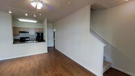 This two-story space is designed with a practical, open layout on the main level. The living area features rich, dark hardwood-style flooring that extends throughout, complemented by clean white walls and white baseboards that create a bright, neutral backdrop. A ceiling fan with a built-in light fixture provides both ventilation and overhead illumination for the main living space.

The kitchen is openly connected to the living area, offering light wood cabinetry, dark countertops, a built-in sink, and black appliances, including a prominent refrigerator. Under-cabinet lighting adds functional brightness to the workspace. A doorway off the main level leads to an additional room or utility space, providing convenient access from the central living area.

A staircase with a white-painted railing runs along one wall, leading to the second floor and adding vertical dimension to the space. The overall design is straightforward and functional, with a cohesive neutral color palette throughout the main level. The open connection between the kitchen and living area supports an easy flow, making the space well-suited for everyday living.
