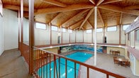 This single-floor amenity space is designed around a striking indoor pool area as its architectural centerpiece. The pool is housed beneath a soaring vaulted ceiling constructed with exposed wood beams arranged in a radial pattern, supported by a central column. Clerestory windows line the upper walls, filling the space with natural light and creating an airy, lodge-inspired atmosphere. A decorative mural wraps around the pool surround, adding a distinctive visual element, while a wraparound upper mezzanine with wood railings offers an elevated view of the pool below.

Adjacent to the pool area, an interior lounge provides a comfortable gathering space furnished with leather sofas, an entertainment center, and dining tables with high-back chairs. Warm-toned walls and carpeted flooring contribute to a relaxed, welcoming ambiance. Interior windows overlook the pool atrium, maintaining a visual connection between spaces.

The communal kitchen is well-appointed with light wood cabinetry, stainless steel appliances including a refrigerator and dishwasher, a long counter-height bar with pendant lighting, and tile flooring. A clean, functional bathroom with a single-sink vanity, light wood cabinetry, mosaic tile flooring, and overhead vanity lighting completes the facility.