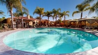 Located at 1510 Santa Diana Rd in Chula Vista, California, this stunning outdoor amenity space showcases a resort-style pool area designed with elegance and relaxation in mind. The centerpiece is a generously sized, freeform swimming pool with pristine turquoise water, surrounded by a spacious pool deck ideal for lounging and entertaining under the Southern California sun.

Tall, mature palm trees are strategically planted throughout the space, lending a tropical ambiance and providing natural shade across the deck. Multiple private cabanas with flowing white curtains are positioned around the pool perimeter, offering shaded retreats with comfortable seating for a premium poolside experience. Additional lounge chairs and patio furniture are arranged along the deck to accommodate guests.

The surrounding architecture reflects a classic Spanish Colonial Revival style, characterized by warm stucco exteriors, red clay tile roofs, decorative arched openings, and wrought iron details. A pergola structure and covered gathering areas further enhance the outdoor living experience.

The overall ambiance is that of a well-appointed community retreat, combining lush landscaping, architectural charm, and thoughtfully designed amenities to create an inviting, resort-like atmosphere perfectly suited to the sunny climate of Chula Vista.